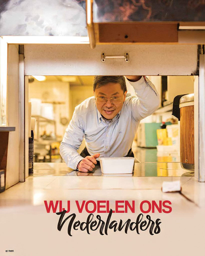 Vriendelijk lachende man met bril leunt door het doorgeefluik van een restaurantkeuken met een wit bakje in zijn hand. Bij het artikel 'Wij voelen ons Nederlanders' in dorpsglossy BAMI.